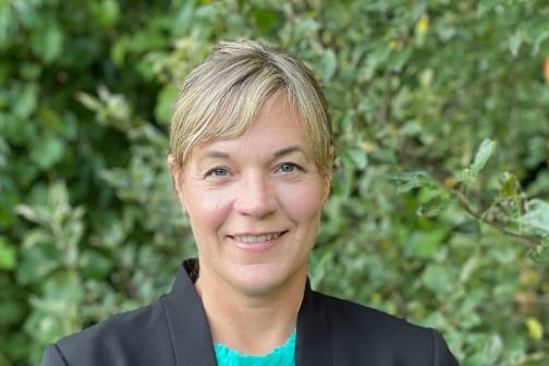 A person with short blonde hair smiles in front of a leafy green background. They are wearing a black blazer and a teal top.