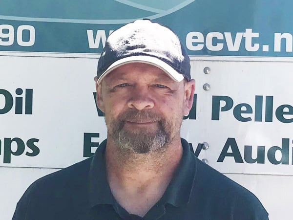 Man with a beard wearing a black baseball cap and dark polo shirt, standing in front of a sign with partial text visible, including Oil and ecvt.